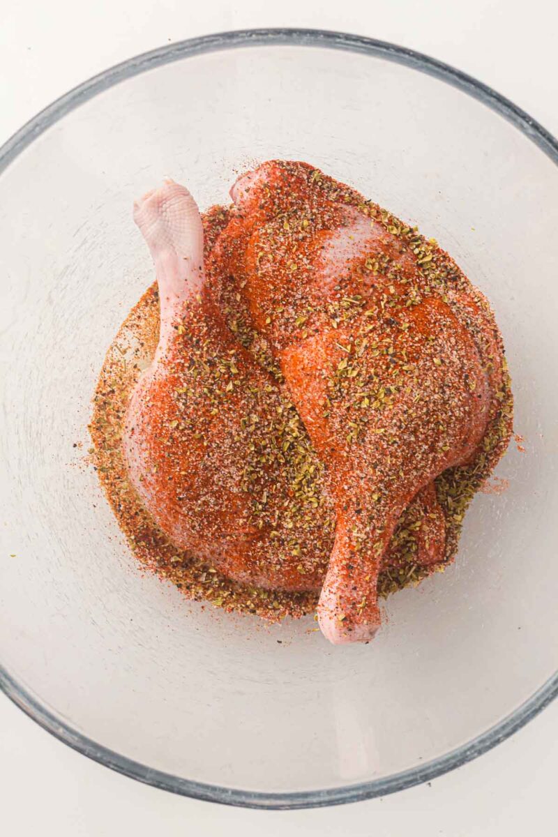 Two raw duck legs coated with a blend of herbs and spices sit in a clear glass bowl.