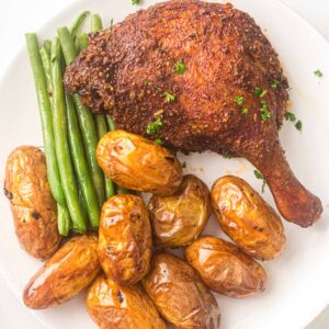 A plate with roasted duck leg, green beans, and roasted baby potatoes on a white background.