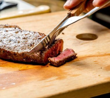 A hand uses a knife and fork to cut a slice from a cooked steak on a wooden cutting board.