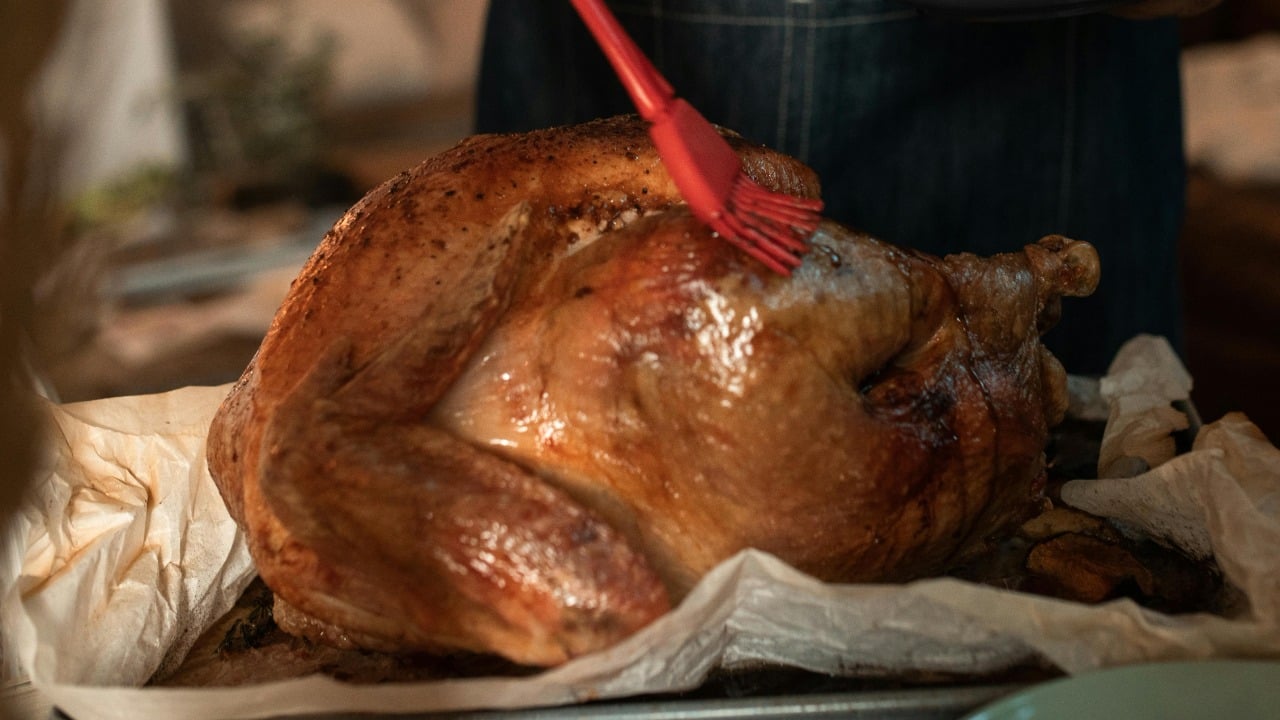 A person uses a red brush to baste a roasted turkey resting on parchment paper in a baking tray.