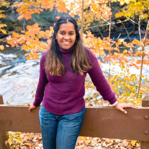 A woman in a purple sweater and jeans leans against a wooden railing with autumn trees and a river in the background.