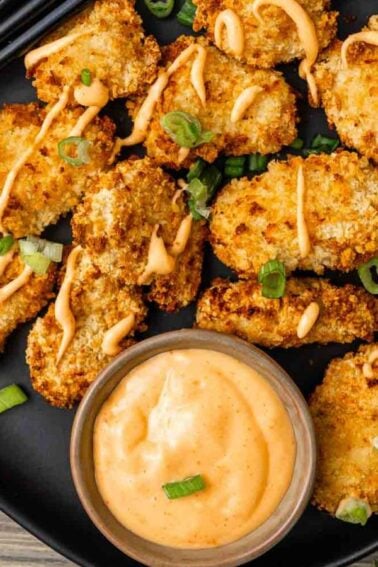 A black plate of breaded, air fryer chicken bites topped with bang bang sauce and chopped green onions, served with a bowl of creamy dipping sauce.