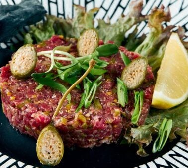 A plate of beef tartare garnished with caper berries, green herbs, and a lemon wedge, served on lettuce with black crackers on the side.