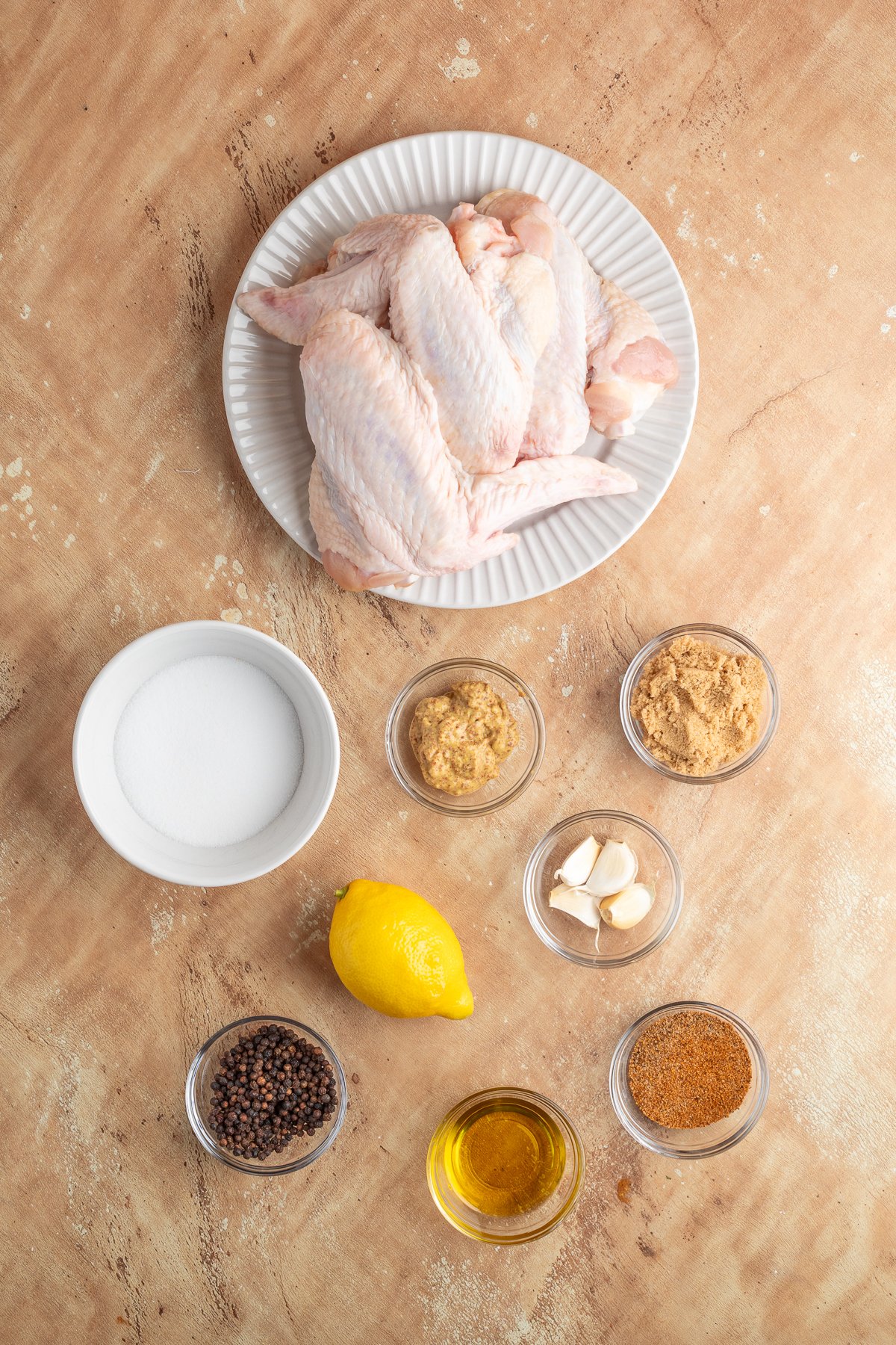 Raw turkey wings on a white plate, surrounded by small bowls of sugar, mustard, brown sugar, garlic cloves, lemon, black peppercorns, olive oil, and seasoning on a beige surface.