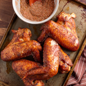 Four seasoned and smoked turkey wings are on a baking sheet next to a bowl of dry spice rub with a wooden spoon.