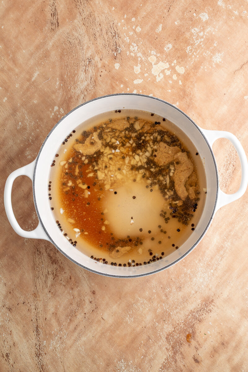 A white pot contains brine with floating minced garlic, black peppercorns, and brown spices on a light wooden surface.