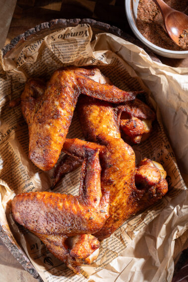Three smoked turkey wings with seasoning, placed on parchment paper over newspaper in a rustic dish, with a bowl of spice mix and wooden spoon nearby.