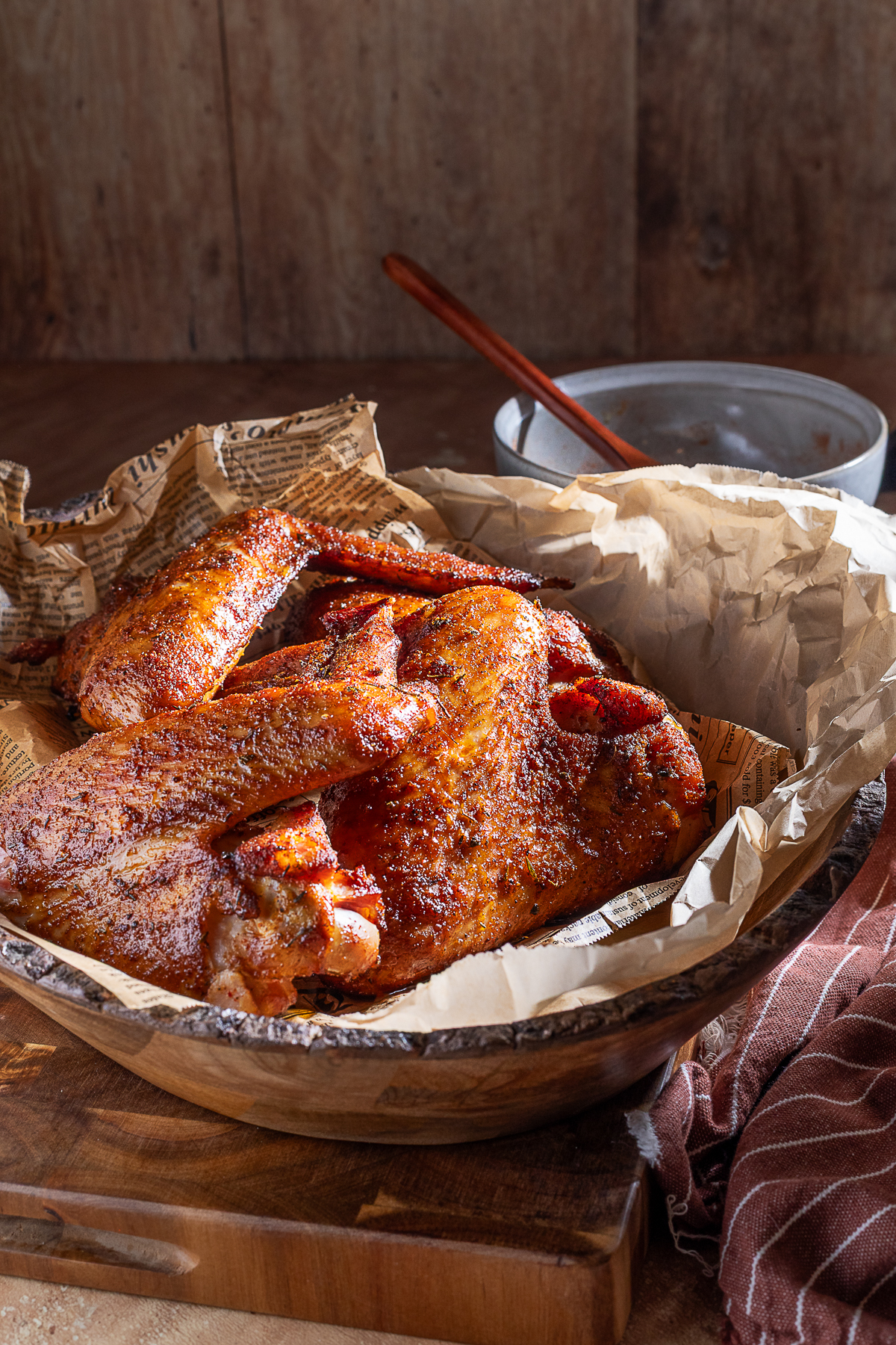 A bowl lined with parchment paper holds several seasoned and smoked turkey wings, with a brown cloth and a glass bowl in the background.