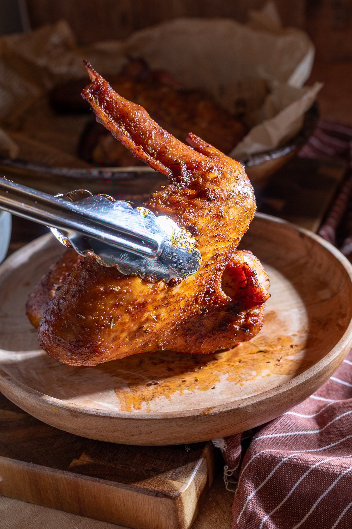 A metal tong holds a cooked, seasoned turkey wing upright on a wooden plate, with a second plate of chicken wings in the background.
