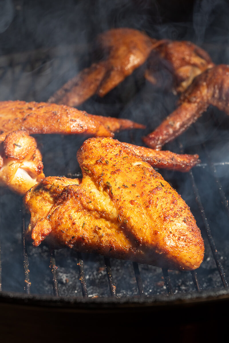 Turkey wings coated in seasoning are being grilled over charcoal, with smoke rising around them.