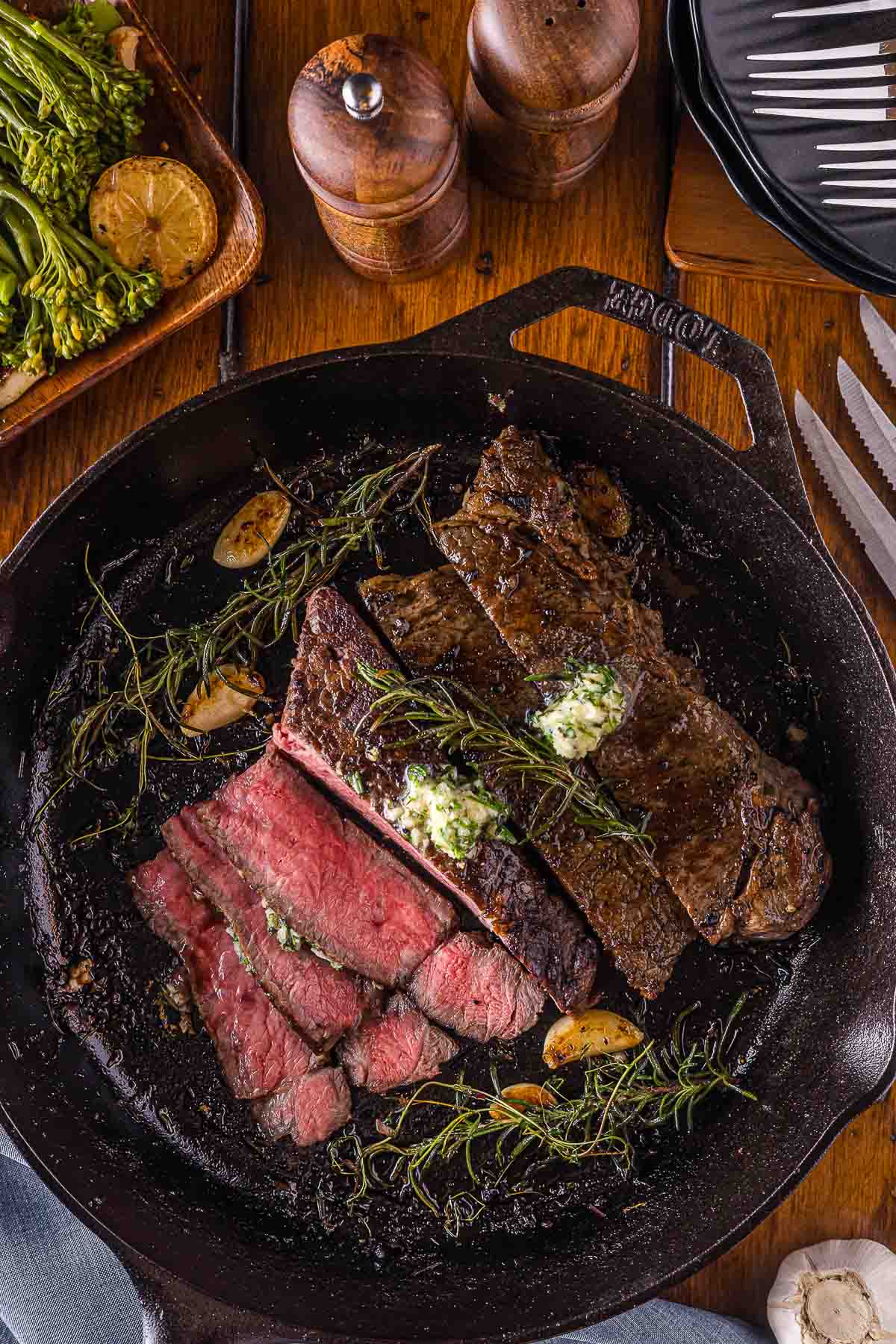 Sliced and whole cooked steak in a cast iron skillet, topped with herb garlic butter and surrounded by garlic cloves and fresh herbs, with vegetables and utensils nearby.