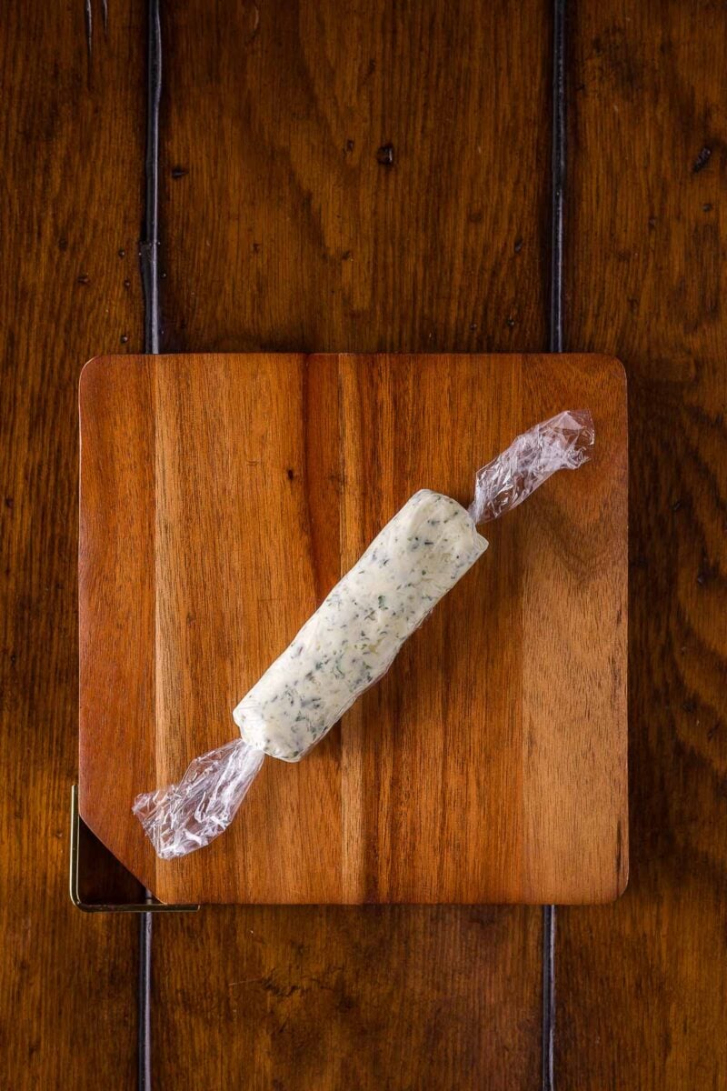 A log of herb butter wrapped in plastic wrap rests on a small wooden cutting board placed on a dark wooden surface.