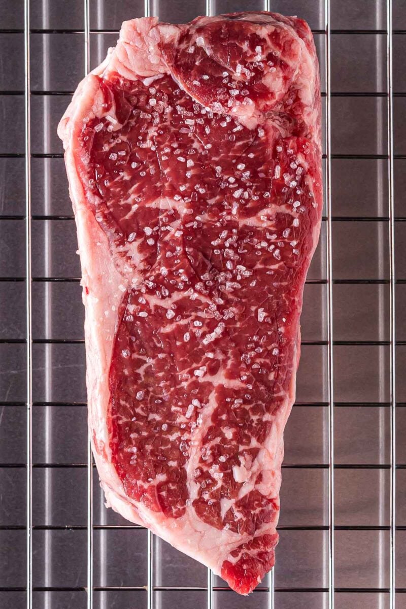 A raw, marbled steak topped with coarse salt sits on a metal wire rack over a grid-patterned surface.