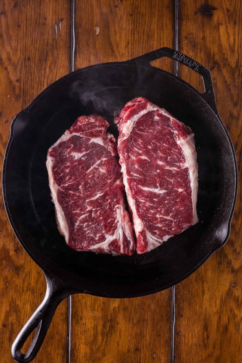 Two raw strip steaks are placed side by side in a black cast iron skillet on a wooden surface.