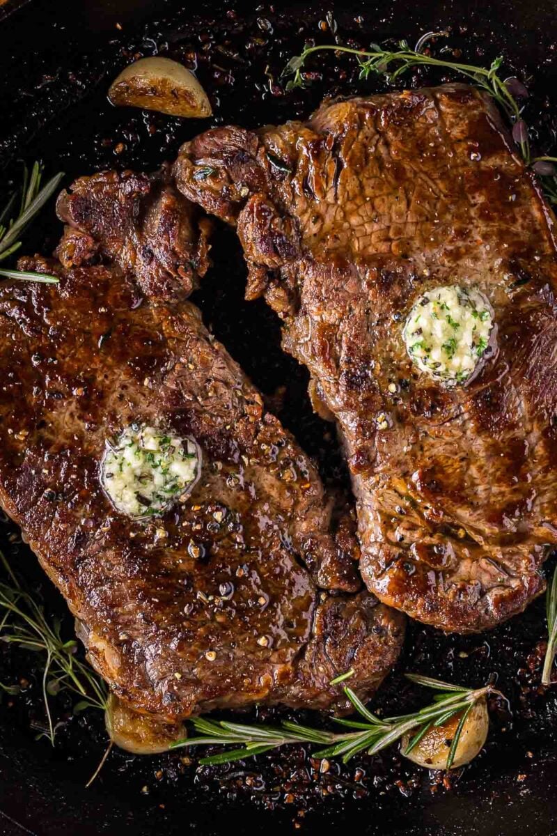 Two cooked steaks in a skillet, topped with herbed garlic butter, surrounded by sprigs of rosemary and garlic cloves.