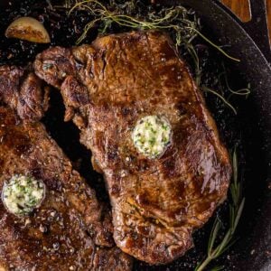 Two cooked steaks in a cast iron skillet with melting herb garlic butter on top, garnished with sprigs of rosemary and roasted garlic cloves.