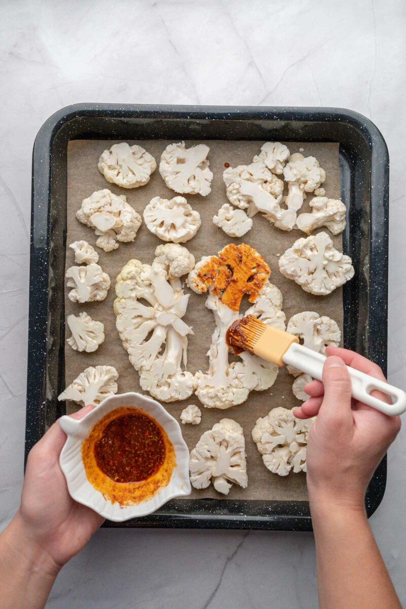 A person brushes seasoning onto raw cauliflower steaks arranged on a parchment-lined baking tray, with a small bowl of marinade nearby.
