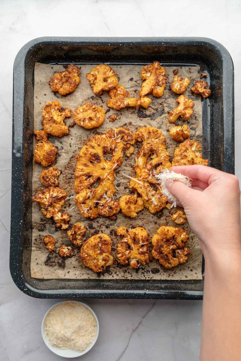 A hand sprinkles shredded cheese over roasted cauliflower steaks and a cauliflower steak on a parchment-lined baking tray.