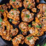 Roasted cauliflower steaks on a black plate, topped with herbs and grated cheese, with lemon wedges and fresh parsley on the side.
