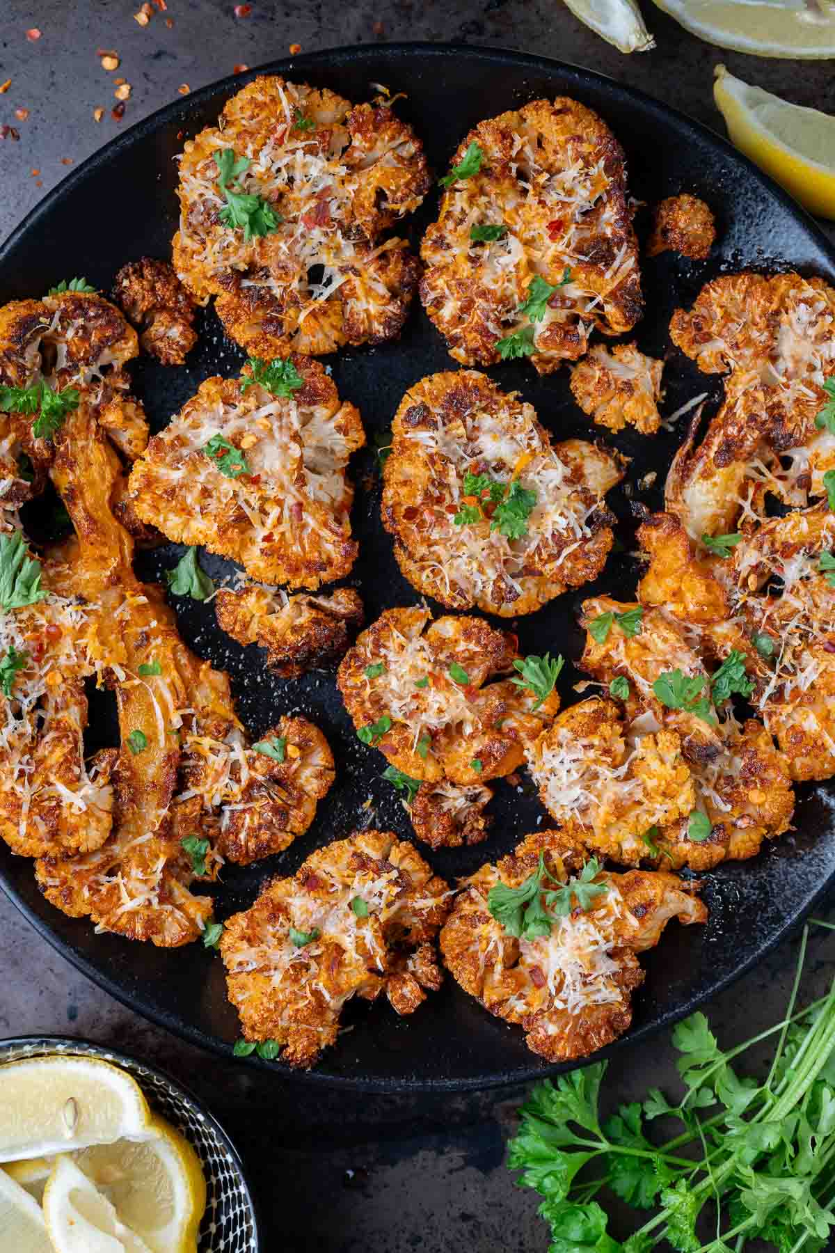 Roasted cauliflower steaks on a black plate, topped with herbs and grated cheese, with lemon wedges and fresh parsley on the side.