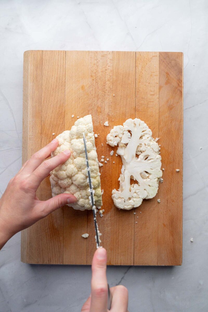 A person slices a head of cauliflower on a wooden cutting board with a large knife.
