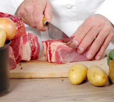 A person in a white coat slices raw beef on a wooden cutting board, surrounded by potatoes, leafy greens, and other vegetables.