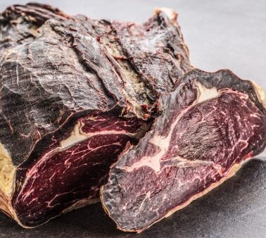 A large piece of dry-aged beef with a dark, crusty exterior and a cut section showing marbled red meat and bone, resting on a gray surface.