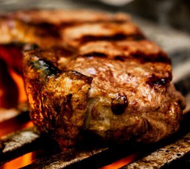 A thick steak is being grilled over an open flame, showing grill marks and a seared surface.