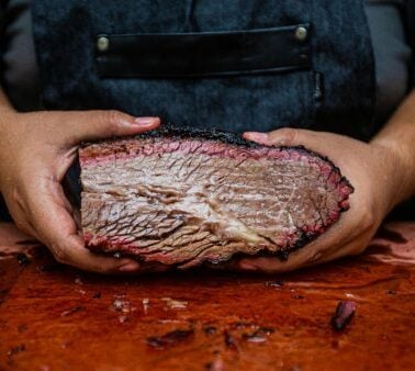 A person holding a large, sliced piece of smoked brisket on a cutting board.