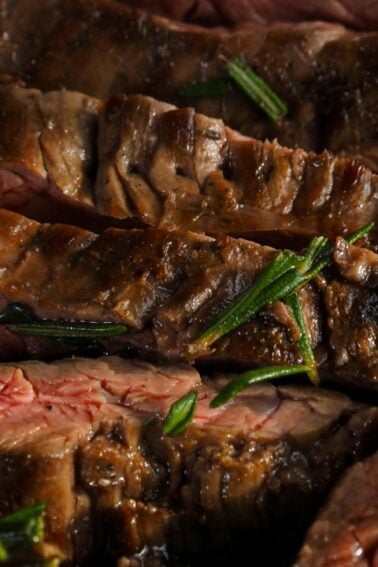 Sliced medium-rare steak with visible grill marks and garnished with fresh rosemary on a wooden cutting board.