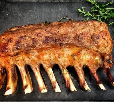 A cooked rack of ribs is placed on a dark surface with a small bunch of fresh herbs beside it.