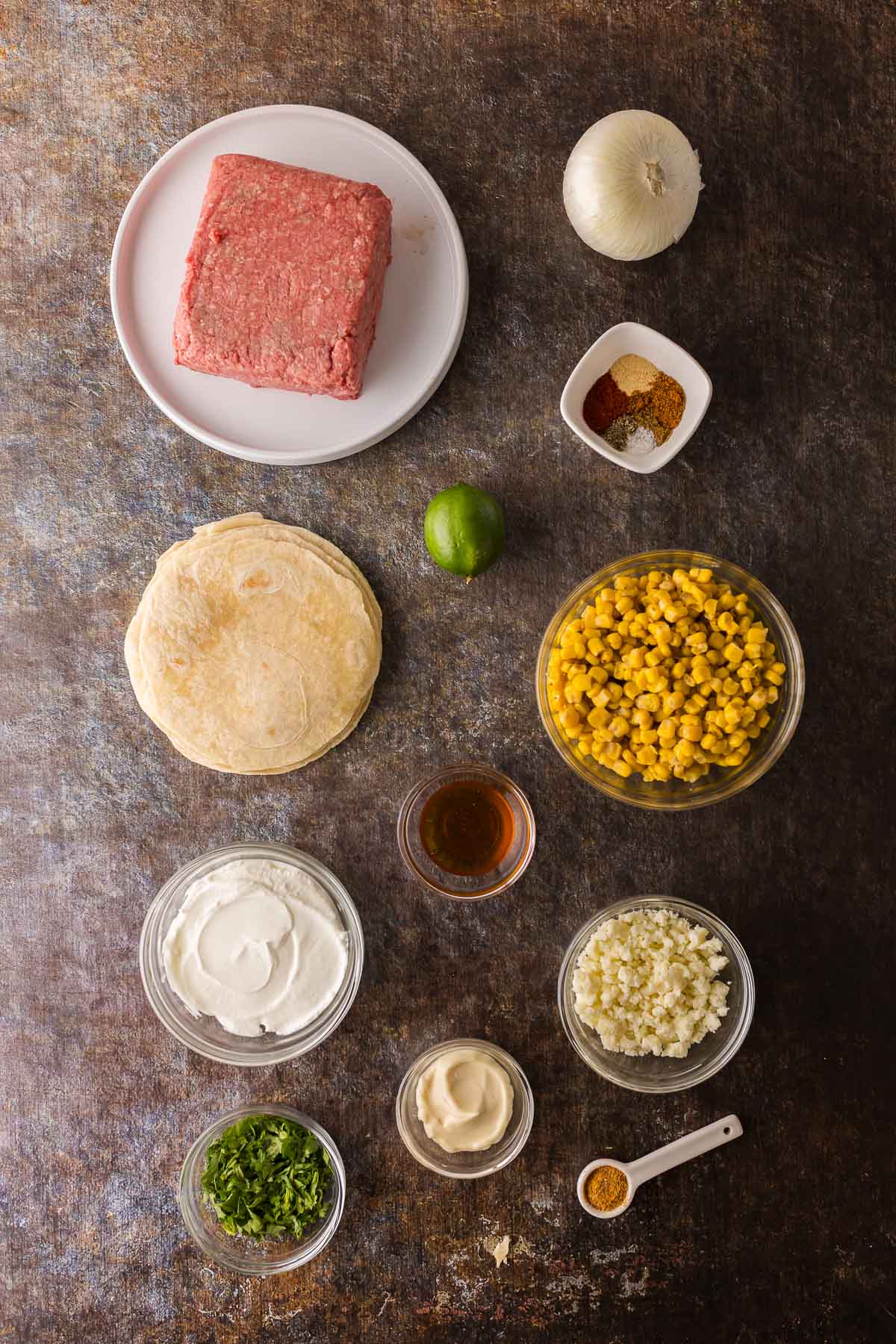 Overhead view of smash taco ingredients including ground beef, tortillas, corn, white onion, lime, sour cream, cheese, mayonnaise, green herbs, spices, and seasoning on a brown surface.