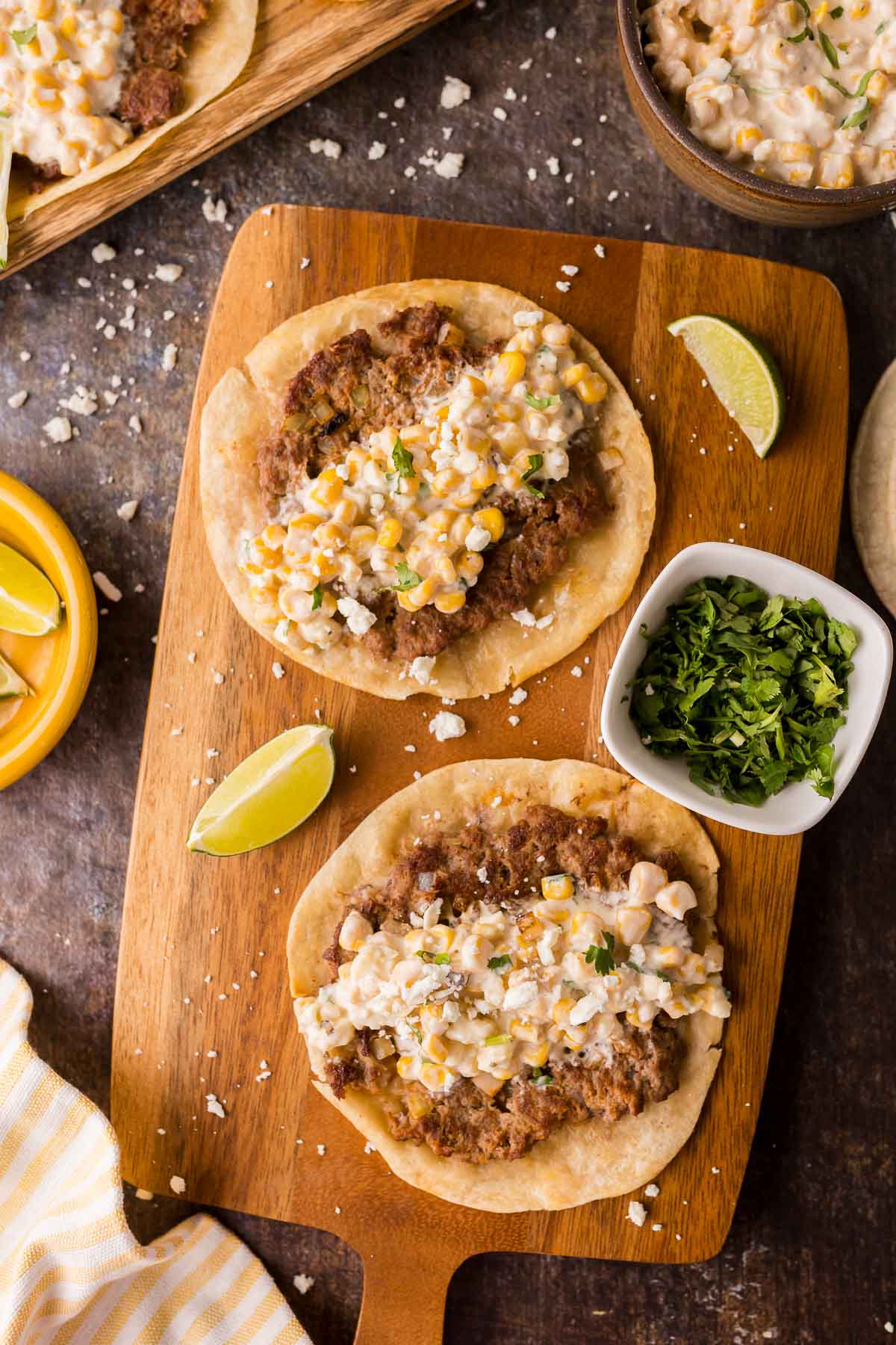 Two smash tacos topped with street corn dip and cheese mixture, garnished with cilantro, served on a wooden board with lime wedges and a small bowl of chopped cilantro.
