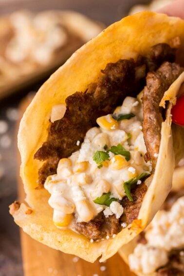 A close-up of a smash taco filled with seasoned beef, creamy corn, chopped onions, cilantro, and crumbled cheese in a soft tortilla.