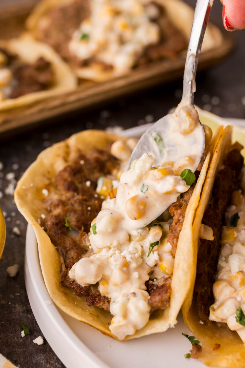 A spoon is spreading creamy street corn dip topping over a taco filled with seasoned ground meat, on a white plate.