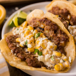 Plate of smash tacos filled with ground meat, creamy corn mixture, and garnished with cilantro, served with lime wedges on the side.