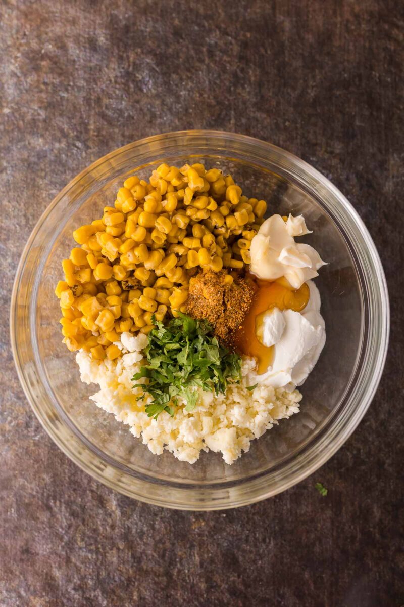A glass bowl containing corn, mayonnaise, sour cream, cheese, spices, and chopped cilantro on a dark surface.