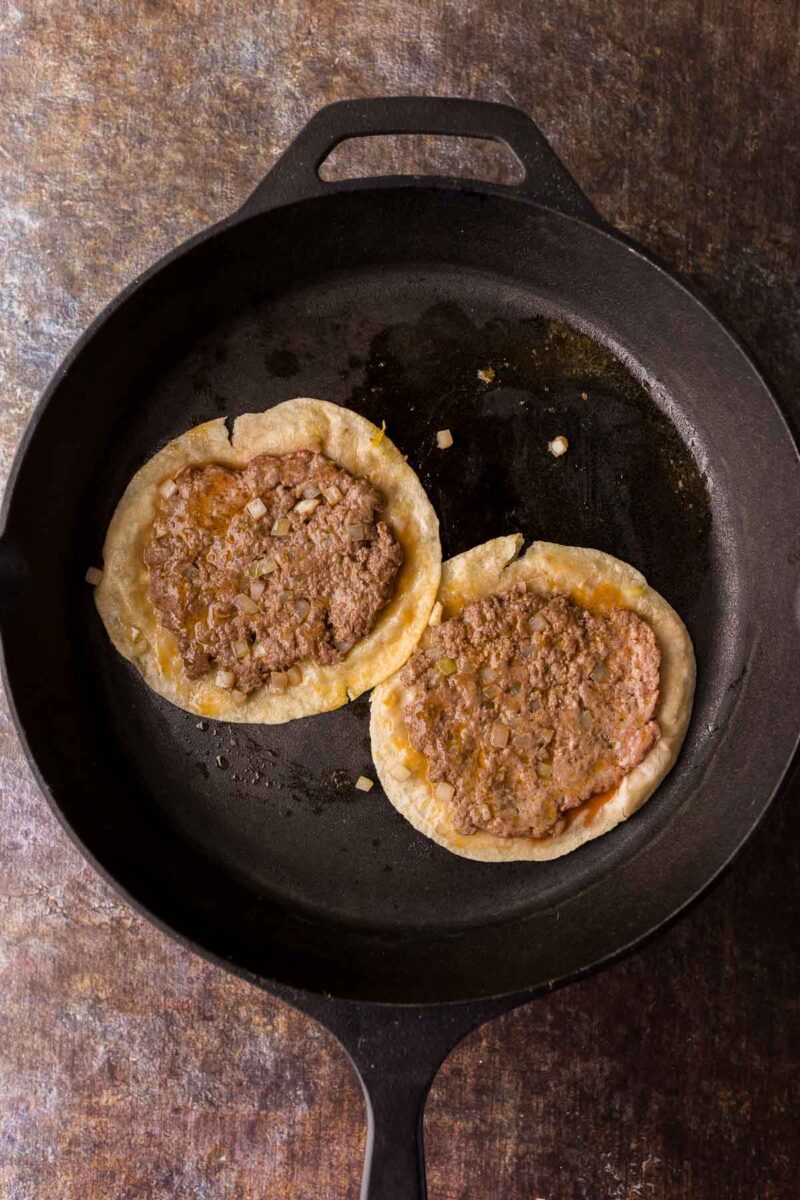 Two small smash tacos are cooking in a black cast iron skillet.
