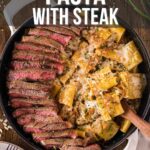 Top-down view of a skillet filled with sliced steak and pasta in a creamy fernch onion pasta, with a wooden spoon resting inside. Text overlay offers a recipe for French onion pasta with steak.