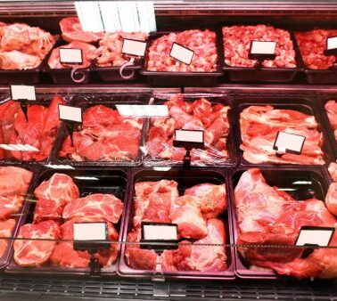 Display case filled with various cuts of raw meat arranged in black trays, each labeled with blank white tags under bright lighting.
