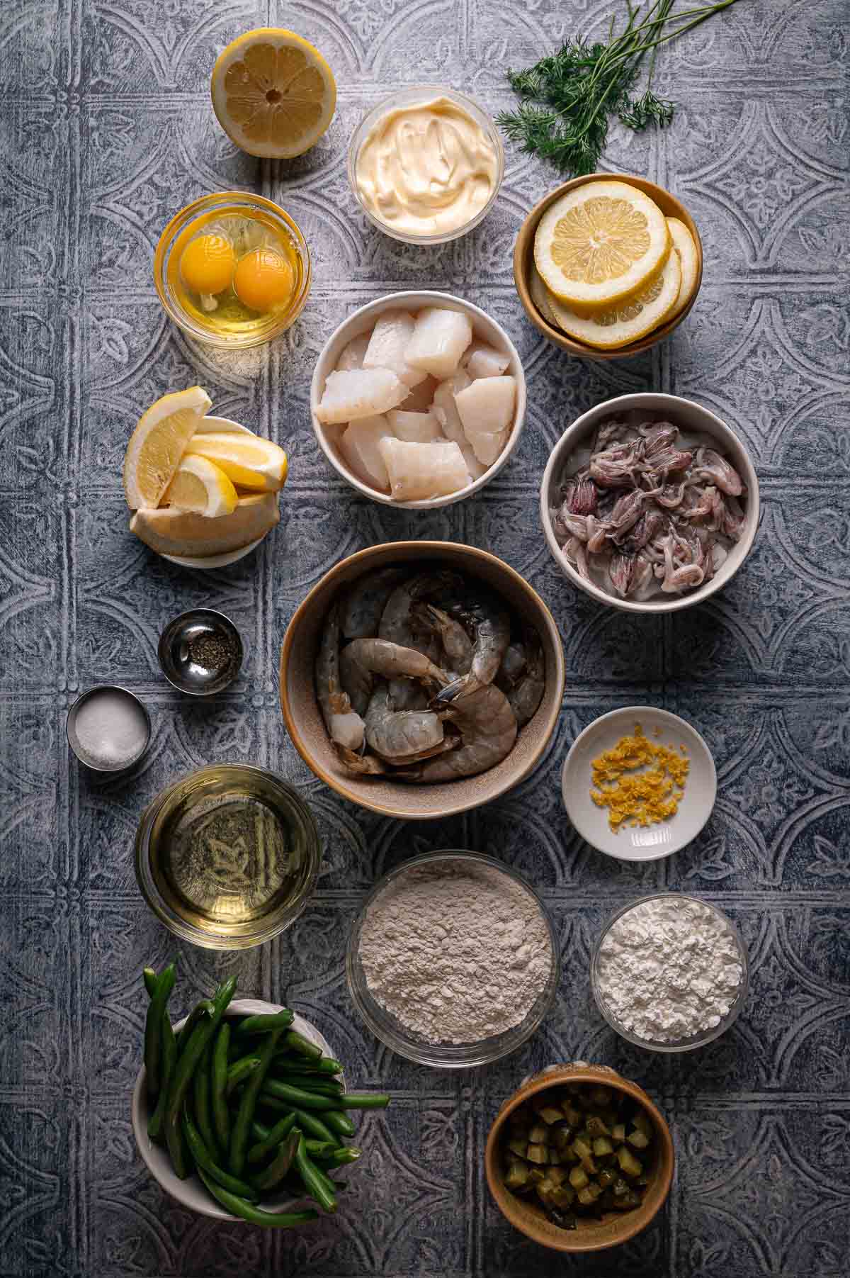 Various ingredients for a fritto misto seafood dish are arranged on a patterned surface, including shrimp, cod, eggs, flour, lemon, green beans, mayonnaise, and seasonings.