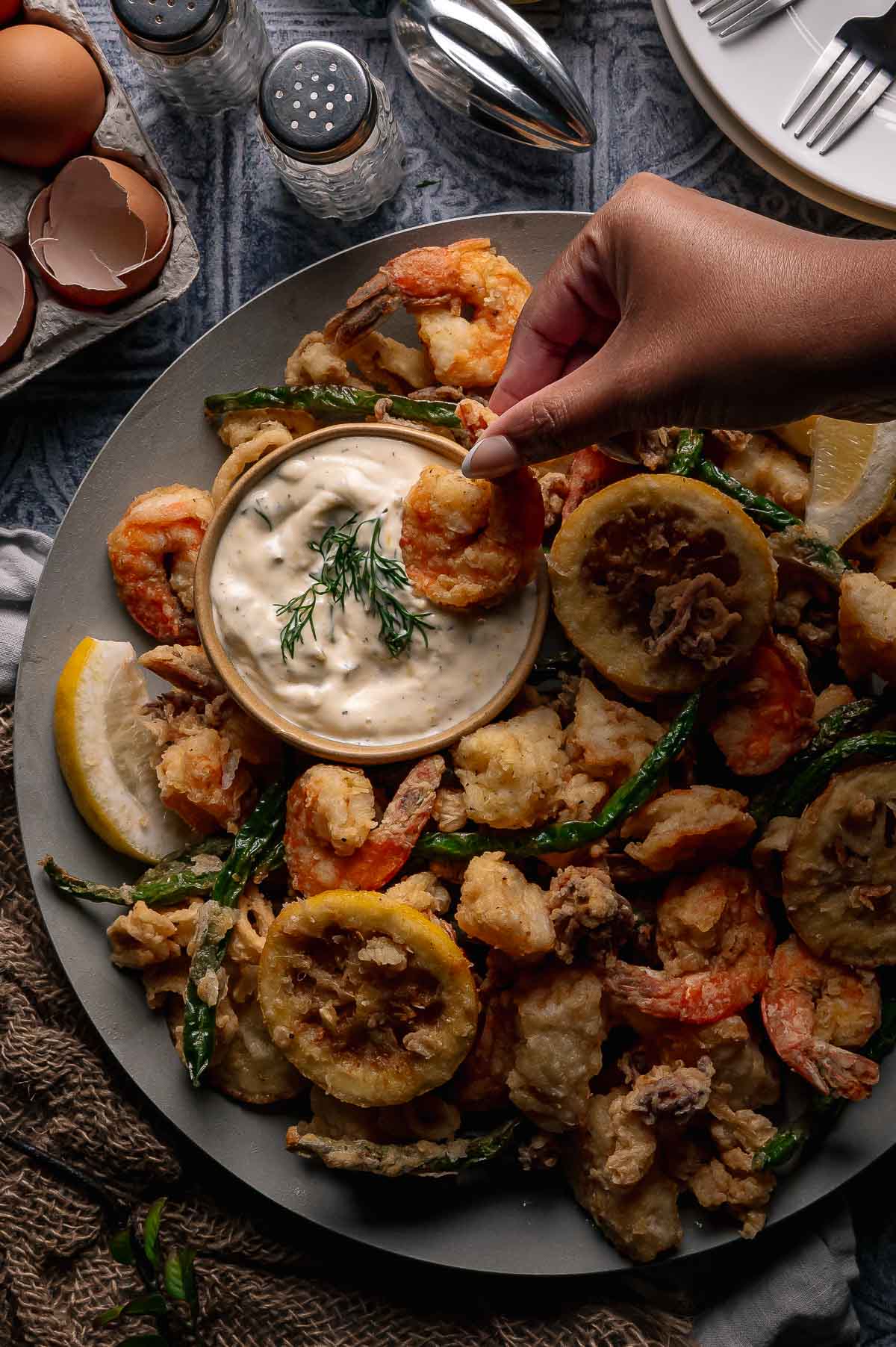 A hand dips a fried shrimp into a bowl of creamy dipping sauce on a platter with fried shrimp, vegetables, lemon slices, and garnishes.