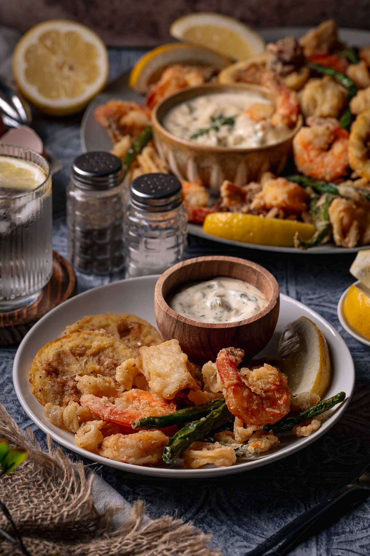 A plate of fritto misto; fried seafood and vegetables with a lemon wedge and dipping sauce, accompanied by a glass of water and condiments.