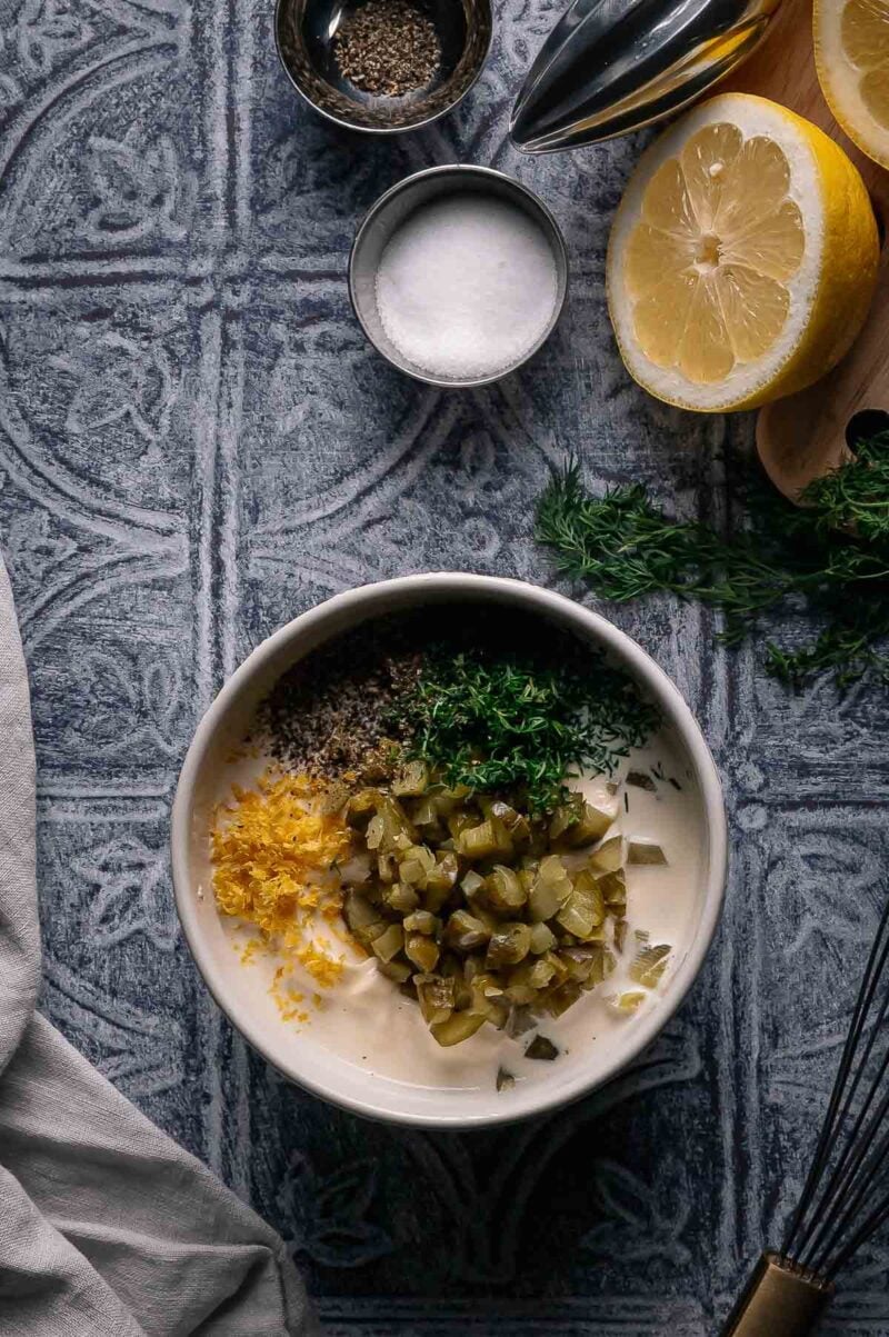 A bowl containing mayonnaise, chopped pickles, lemon zest, fresh herbs, ground pepper, and salt, with half a lemon, salt, pepper, and fresh herbs on the side.
