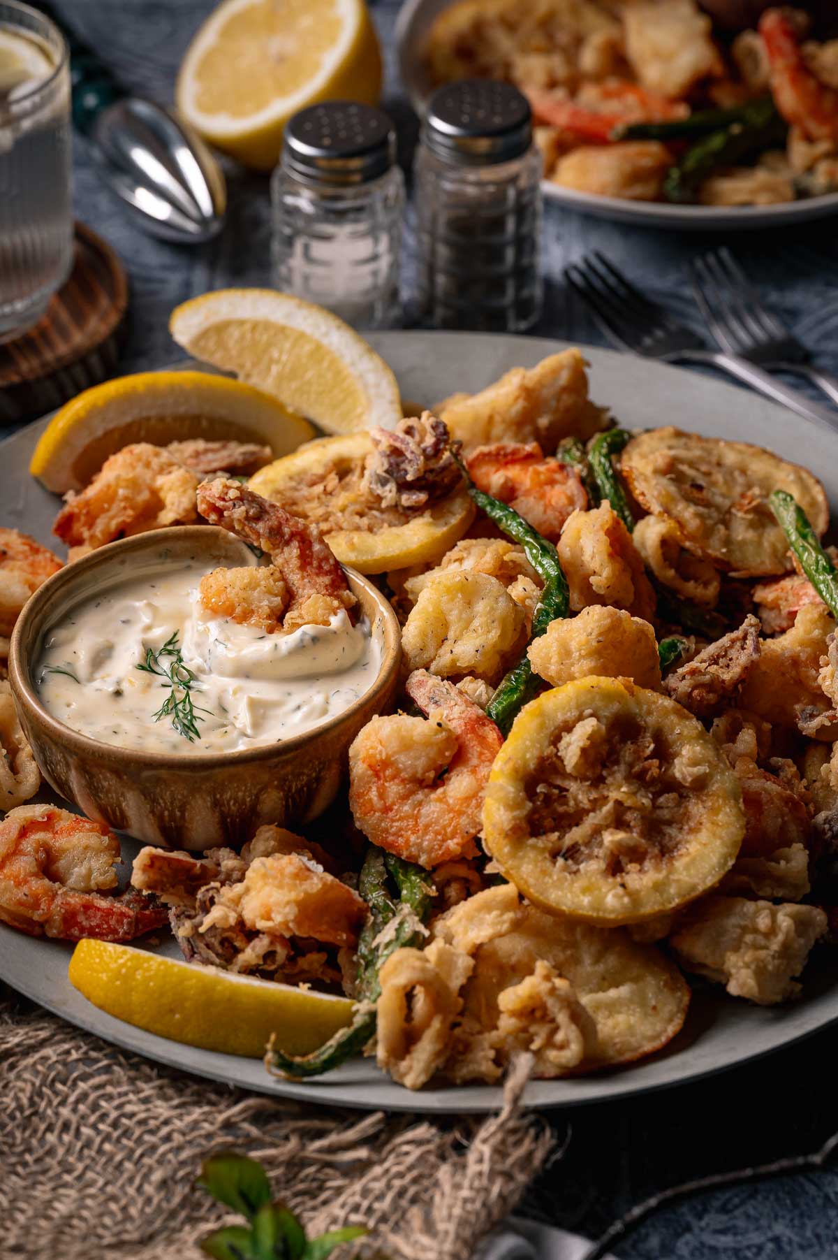 A plate of fritto misto; assorted fried seafood and vegetables with lemon wedges and a bowl of creamy dipping sauce, garnished with dill.