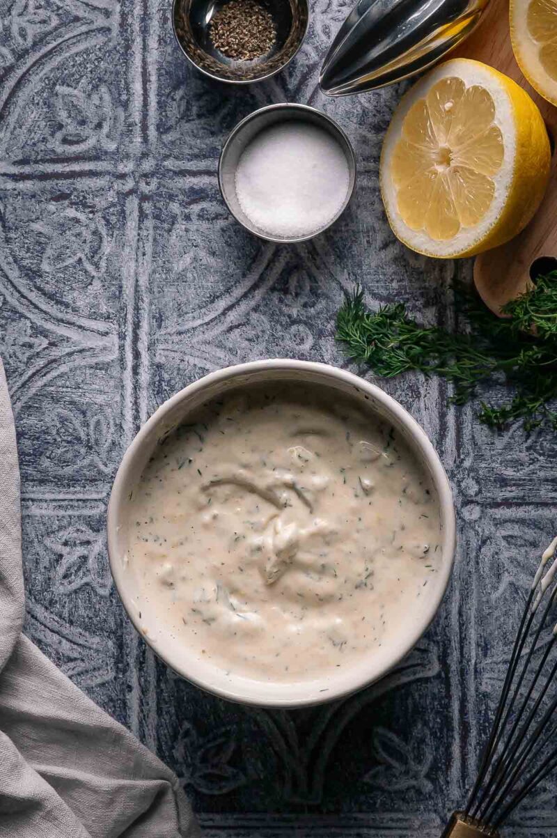Bowl of creamy lemon dill tartar sauce with herbs, surrounded by a halved lemon, salt, pepper, fresh dill, and a whisk on a patterned surface.