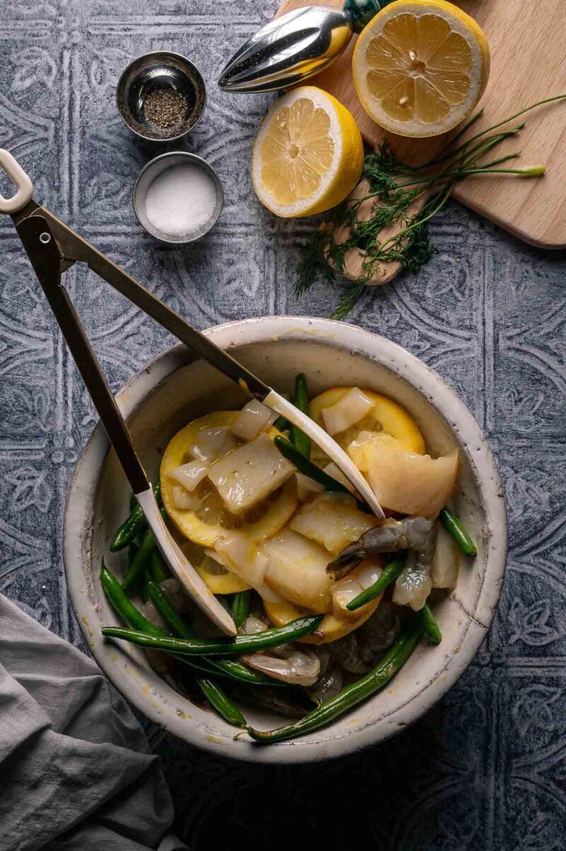 A bowl with raw shrimp, lemon slices, cod, and green beans, with tongs resting on top. Halved lemon, salt, pepper, and herbs are nearby on a textured surface.