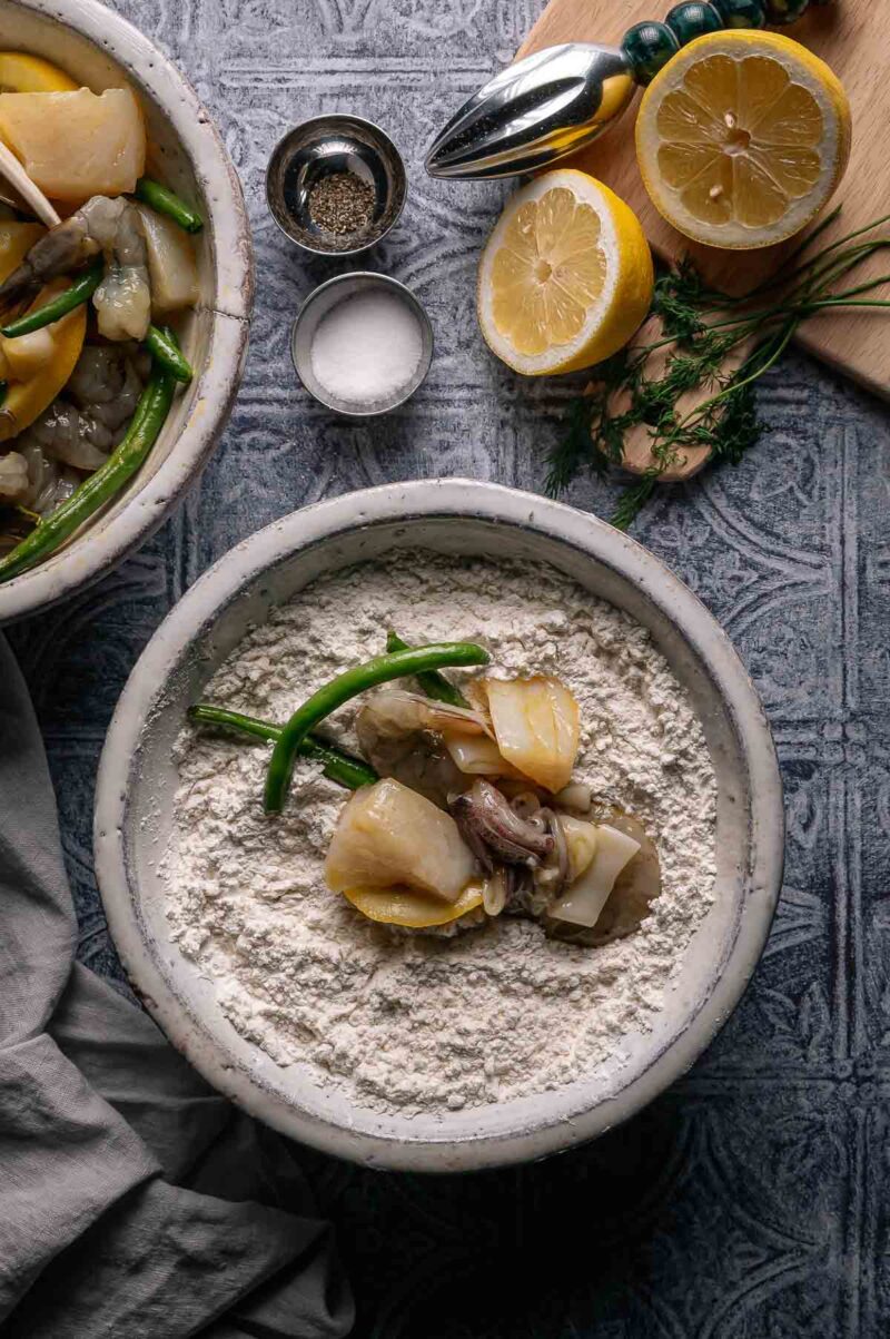 A bowl with flour, raw shrimp, cod, and green beans. Nearby are lemon halves, salt, pepper, and fresh herbs on a cutting board.