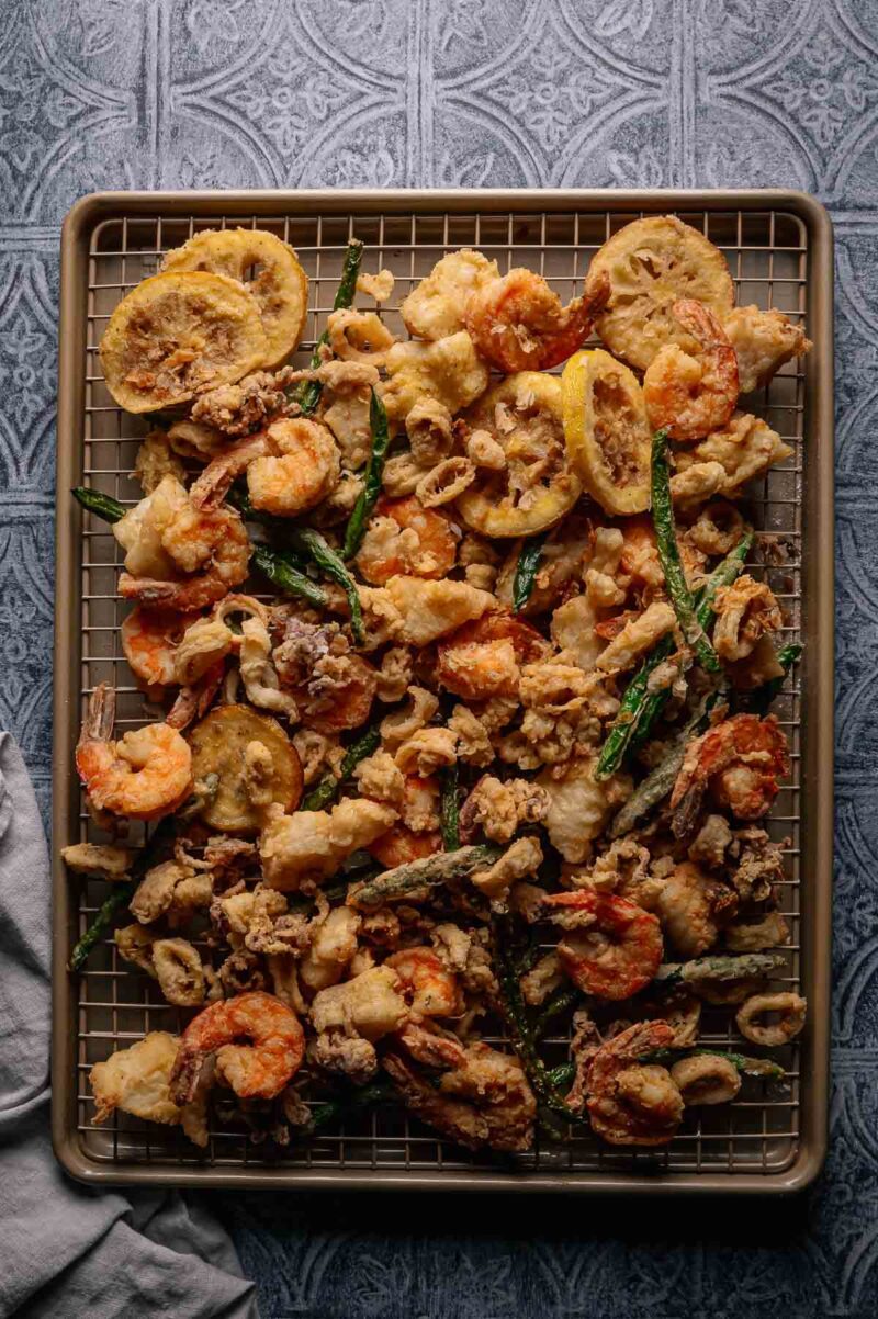 A tray of assorted fried seafood, fritto misto, including shrimp, calamari, and vegetables like lemon slices and green beans, arranged on a wire rack over a baking sheet.