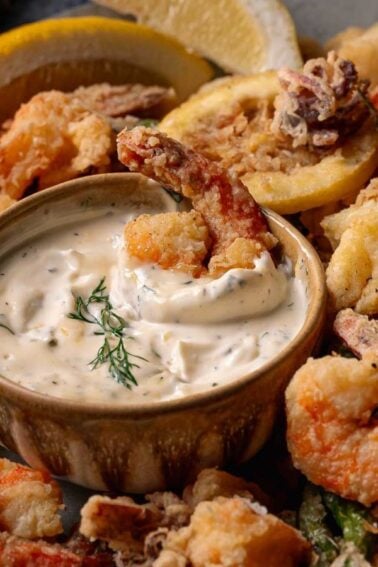 A plate of fritto misto, fried seafood and vegetables with a bowl of creamy dipping sauce in the center, garnished with lemon wedges.